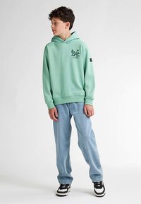 Mintgrön hoodie med en känguruficka, ribbade muddar och en logografik. Kombinerad med ljusblå jeans och svartvita sneakers.