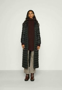 Lang svart pufferjakke med horisontal quilting, båret over en mørk halskjorte, kombinert med grå mønstrede bukser og blanke brune støvler.