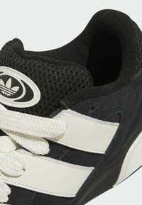 Mustat samettiset tennarit valkoisilla verkko- ja nahkaosuilla, joissa on paksut valkoiset nauhat ja pyöristetty kärki, esittelee Adidas-logoa.