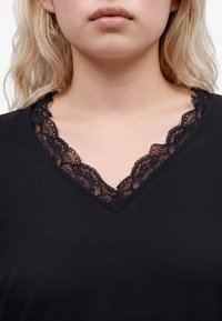 Gros plan d'une personne portant un haut noir à col en V avec une bordure en dentelle noire le long de l'encolure et cheveux blonds partiellement visibles.