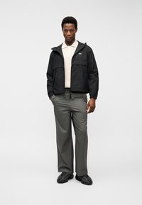 Άνδρας που φορά μαύρο μπουφάν με κουκούλα, λευκό πόλο μπλουζάκι, γκρίζο παντελόνι και μαύρα αθλητικά παπούτσια, στέκεται με τα χέρια στις τσέπες πάνω σε λευκό φόντο.