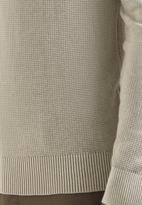 Close-up van een beige wafelsteek trui met geribde mouwen en onderkant, gedragen over een bruine broek.