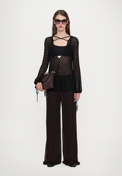 Cardigan nero in maglia trasparente sopra una maglietta nera, abbinato a pantaloni larghi con motivo. La modella tiene una borsa bordeaux e indossa occhiali da sole.