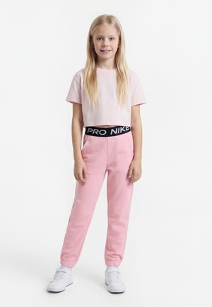 Blond jente står, iført en lys rosa cropped t-skjorte, rosa joggebukser med svart Nike Pro linning og hvite sneakers.