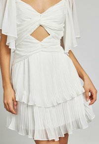 Weißes plissiertes Kleid mit fließenden Ärmeln, das über einen verdrehten Oberteil und eine ausgeschnittene Mitte verfügt. Geschichtete Stufen sorgen für Textur und Bewegung.