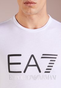 Biała bawełniana koszulka z dużym czarnym nadrukiem w postaci logo "EA7 EMPORIO ARMANI" z przodu, okrągły dekolt o gładkiej fakturze.