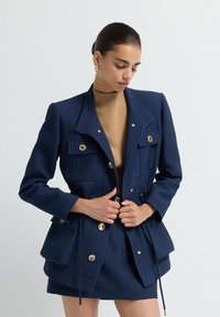 Kobieta poprawiająca granatową, teksturowaną marynarkę z złotymi guzikami, w zestawie z pasującą spódnicą i camelowym golfem na tle gładkiej białej ściany.