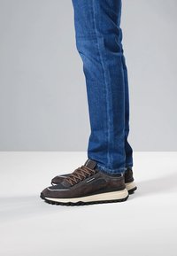 Sneakers in camoscio marrone scuro con lacci marroni, dettagli bianchi e una suola spessa in gomma nera, abbinati a jeans blu.