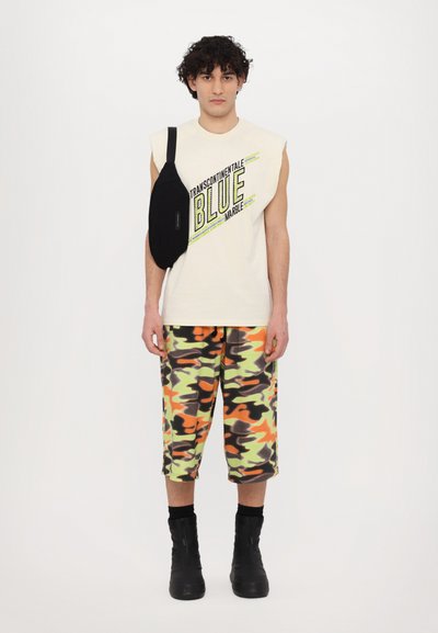 Kremfarget ermeløs skjorte med "BLUE MARBLE" trykk, paret med camo-shorts i oransje, grønn og svart. Svart crossbody-veske og svarte støvler.