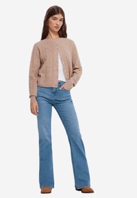 Beige gebreide cardigan met een subtiele textuurpatroon, knoopsluiting en geribbelde manchetten, gecombineerd met lichtblauwe flared jeans en een paar bruine laarzen.