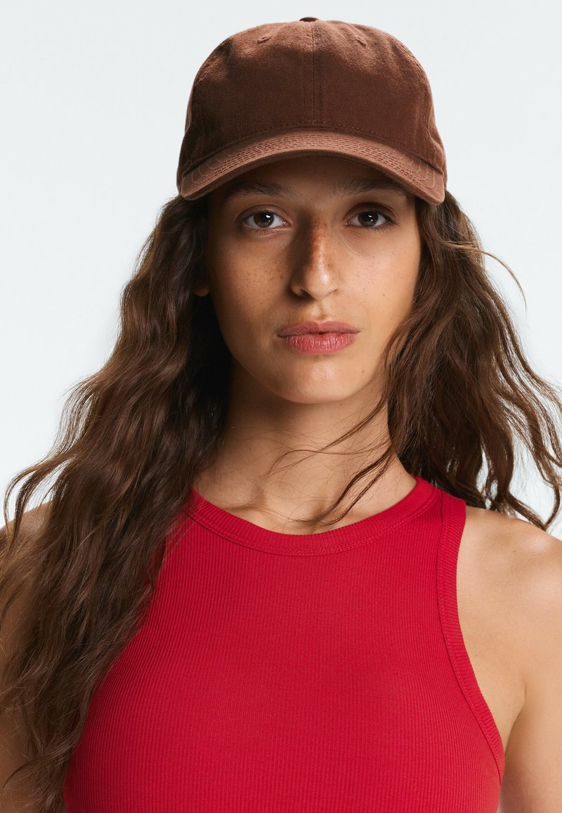 Jeune femme aux longs cheveux ondulés portant une casquette marron et un haut rouge sans manches, regardant droit devant elle avec une expression neutre.
