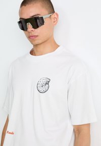 Young man wearing large angular black sunglasses and a white T-shirt with a circular black graphic and "Fossils" text.