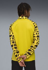 Gelbes Sportshirt mit schwarzen geometrischen Mustern an den Ärmeln. Hat eine glatte Textur und ein gerades Schnittdesign.