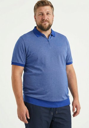 Bärtiger Mann steht, trägt ein blaues kurzärmeliges Polohemd mit Reißverschluss und dunkle Hose vor hellem, einfarbigem Hintergrund.