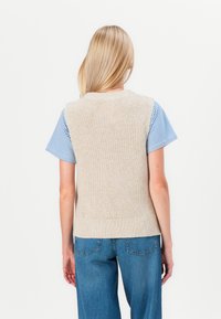 Gilet écru en maille sans manches avec un motif à côtes et un col, associé à une chemise à rayures bleues et blanches à manches courtes et un pantalon en denim.