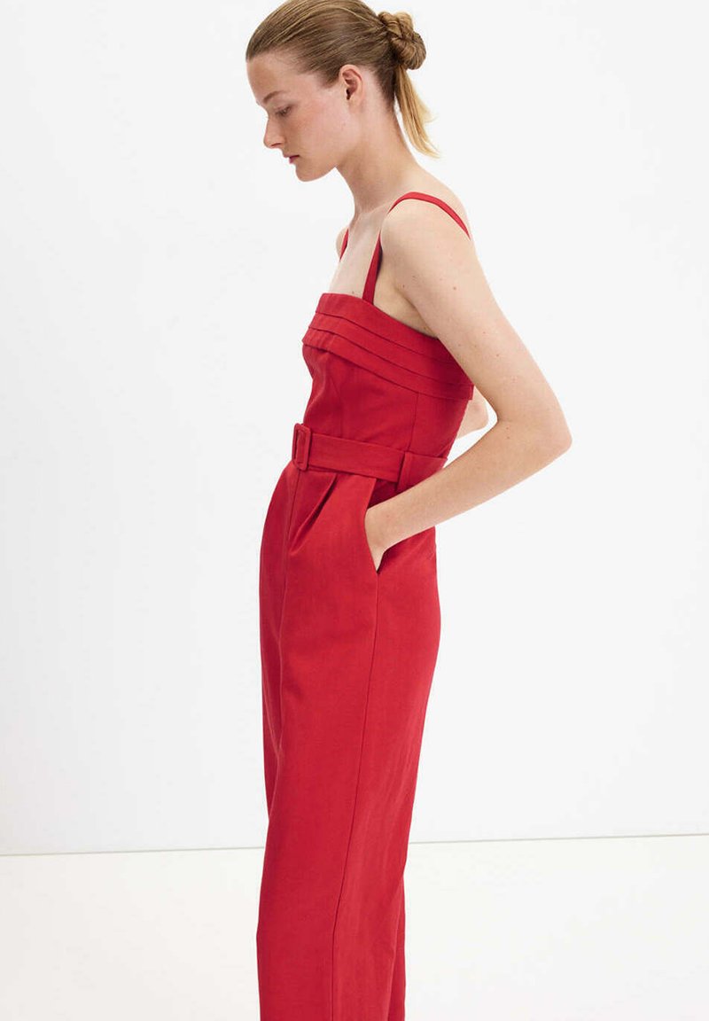 Femme aux cheveux en chignon portant une robe rouge sans manches ceinturée avec un corsage plissé, debout de profil les mains dans les poches sur fond blanc.