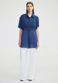 Camisa de manga corta azul marino con cuello, que presenta un panel más claro y bordado estampado en el pecho, combinada con pantalones de pierna ancha blancos.