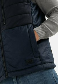 Námořnická puffer vesta s texturovanou tkaninou v horní části a hladkým materiálem dole. Obsahuje zip, žebrové šedé manžety a nášivku s logem.