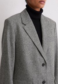 Blazer gris en mélange de laine avec revers crantés, deux boutons et une poche poitrine. Porté sur un col roulé noir côtelé.