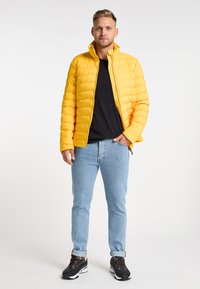 Geel gewatteerd jack met hoge kraag, gequiltde textuur, zwart T-shirt eronder, lichtblauwe jeans en zwarte sneakers met rode accenten.