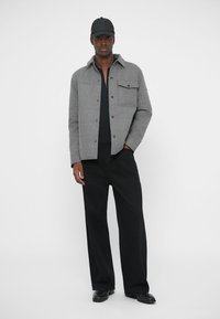 Šedá texturovaná bunda s knoflíkovým zapínáním, dvěma předními kapsami, vrstvená přes černý top. V kombinaci s černými širokými kalhotami a černou čepicí.