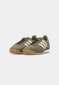 Green athletic shoes with white stripes, made of mesh and suede. Features a gum rubber outsole and brown laces. Classic retro design.