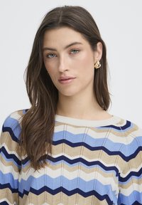 Pull en maille avec un motif en vagues bleu, beige et blanc, avec un col côtelé et un tissu texturé. Le modèle porte des boucles d'oreilles dorées.