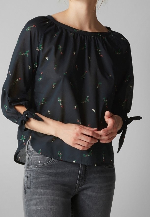 Femme portant un chemisier noir, ample, avec de petits imprimés floraux, manches 3/4 nouées aux poignets, et un jean en denim gris.