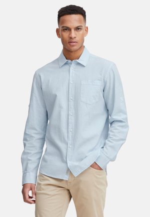 Light blue, long-sleeve button-up shirt in a textured fabric with a chest pocket and rolled cuffs, paired with beige pants.