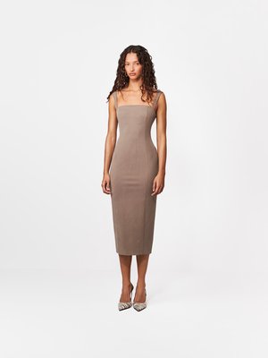 Kobieta stojąca, ubrana w dopasowaną bezrękawnikową sukienkę midi koloru taupe oraz czarno-białe paskowane szpilki na tle jednolitego białego tła.