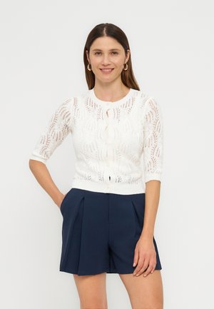 Femme aux longs cheveux bruns portant des boucles d'oreilles créoles dorées, un cardigan blanc en dentelle boutonné, et un short plissé bleu marine, debout la main dans la poche.