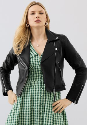 Frau mit langen blonden Haaren trägt silberne Creolen, ein grünes kariertes Hemdkleid und eine schwarze Lederjacke mit Reißverschluss-Details.