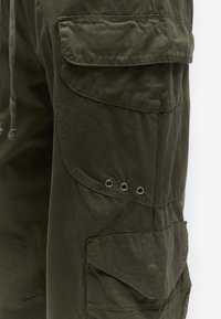 Close-up of olive green cargo pants with multiple flap pockets and metal eyelet vents on the side thigh area.