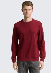 Felpa a maniche lunghe color bordeaux con tessuto testurizzato, scollo rotondo e polsini a coste. Presenta una toppa con logo sulla manica sinistra.