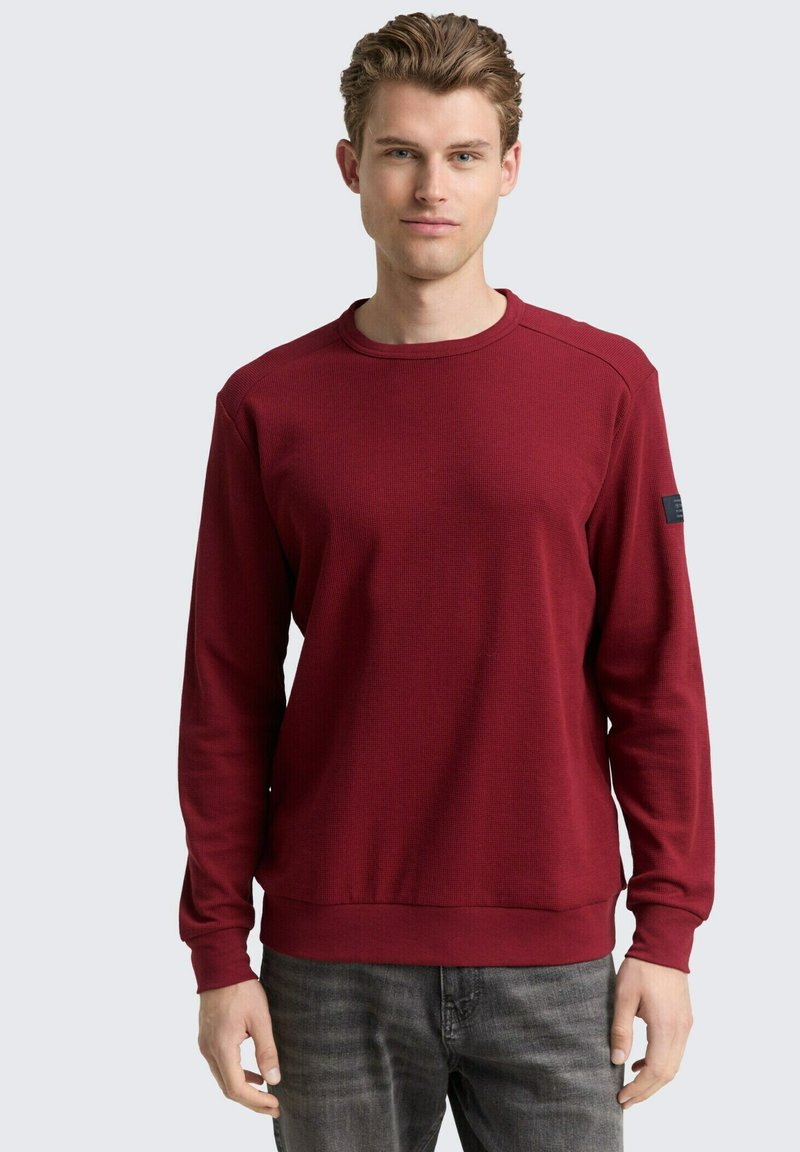 Felpa a maniche lunghe color bordeaux con tessuto testurizzato, scollo rotondo e polsini a coste. Presenta una toppa con logo sulla manica sinistra.