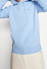 Felpa di cotone azzurro con tasca frontale, cappuccio con cordini e piccolo logo sul petto. Texture morbida con polsini e orlo a coste.