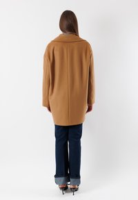 Cappotto di lana marrone chiaro con colletto ampio, spalle scese e una vestibilità rilassata. Abbinato a jeans a zampa d'elefante blu scuro e tacchi neri.