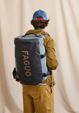 Person wearing mustard jacket and shorts, blue and red cap, carrying large black and gray backpack with "FAGUO" logo on back.