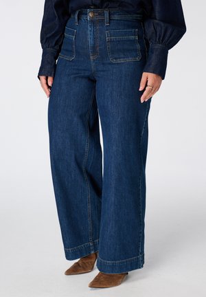 Femme portant un jean bleu foncé taille haute, jambes larges, avec de grandes poches avant et des chaussures en daim marron à bout pointu.