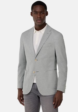 Blazer texturizado gris claro con solapas de muesca, dos bolsillos frontales, cierre de un solo botón y un sutil patrón de malla; combinado con una camiseta polo color crema.