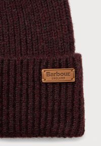 Tumedast punakaspruun ribaga kootud beanie müts, mille kokkuvolditud ääres on pruun nahast silt "Barbour England", valge taustal.