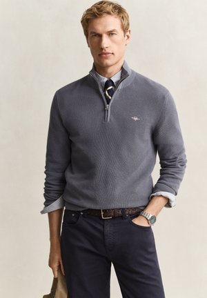 Young man wearing a gray quarter-zip sweater over a shirt and tie, dark pants, a silver wristwatch, and holding a beige hat.