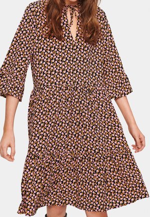 Kobieta w czarnej sukience do kolan z różowo-żółtym kwiatowym wzorem, z rękawami 3/4 oraz dekoltem w kształcie łezki z wiązaniem.