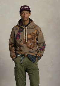 Hanoracul cu glugă are un design cu carouri maronii și imagini imprimate mari și colorate. Este asortat cu pantaloni cargo măslinii și o șapcă roșu-maroniu.