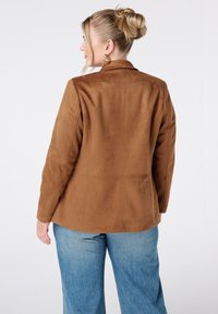 Blazer en suède synthétique marron avec un design structuré, col cranté et détails de coutures visibles. Assorti à un jean large bleu clair.