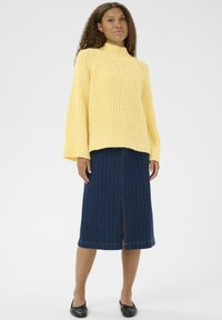 Femme aux cheveux bouclés portant un pull en tricot jaune, une jupe midi en denim bleue avec une fente à l'avant, et des ballerines noires, se tenant sur un fond blanc.