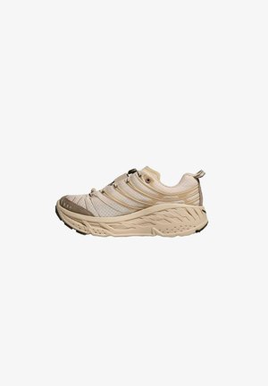 Zapatilla deportiva beige con parte superior de malla texturizada, puntera reforzada, suela acolchada y sistema de cordones integrado. Presenta una banda de rodadura distintiva y acentos.