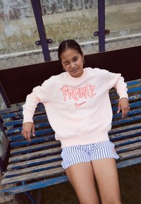 Sudadera rosa claro con la palabra "Francia" en escritura coral y el logo de Nike, combinada con pantalones cortos a rayas azules y blancos, sentado en un banco de madera.