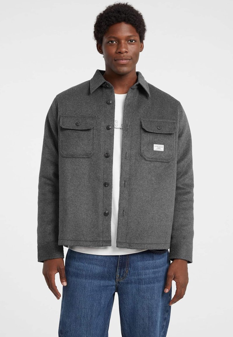 Giacca camicia in lana grigia con due tasche sul petto, chiusura a bottoni e orlo dritto. Indossata sopra una camicia bianca e jeans blu.
