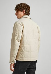 Beige quilted jacket with a structured design, buttoned cuff details, and a classic collar. Worn over dark jeans.